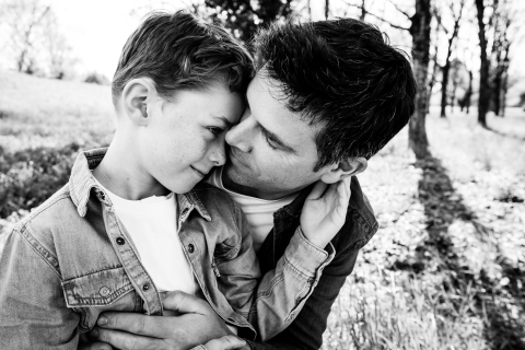 In a black and white photo taken in Miribel, France, a boy and his dad share a warm hug outdoors, surrounded by the beauty of nature, capturing a special father-son scene in a classic style.