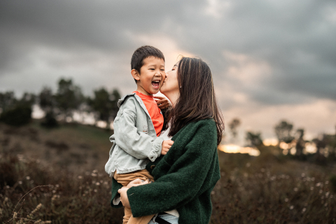 A picture taken during a photo shoot in Los Angeles, California shows a boy smiling happily while his mom gives him a kiss on the cheek, set against a beautiful field of green grass.