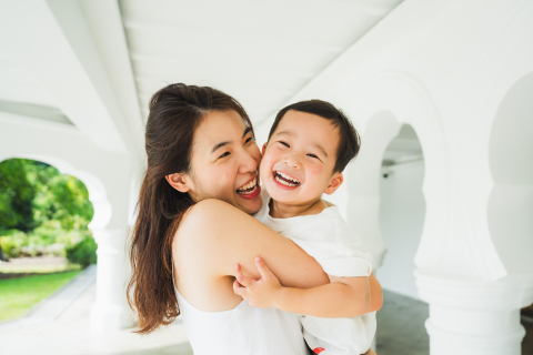 A lifestyle image created at The Gallop Extension of the Botanic Gardens, Singapore captures a special hug as a mother lovingly embraces her son, both smiling happily on a clean white porch.