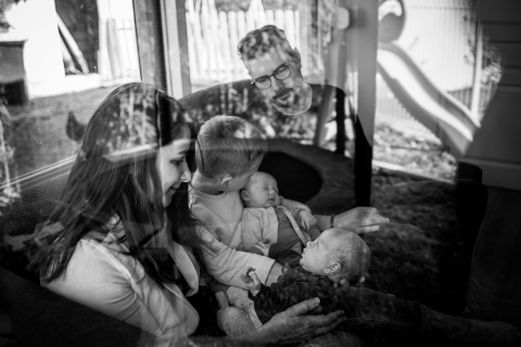 The picture shows a family enjoying time together at home in Montpellier, France, with the focus on a parent holding a baby, captured in black and white through glass.