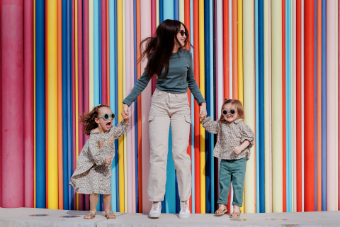 Em Paris, três irmãs usam óculos escuros e se divertem dançando e brincando juntas em frente a uma parede pintada e brilhante durante uma sessão de fotos de família.