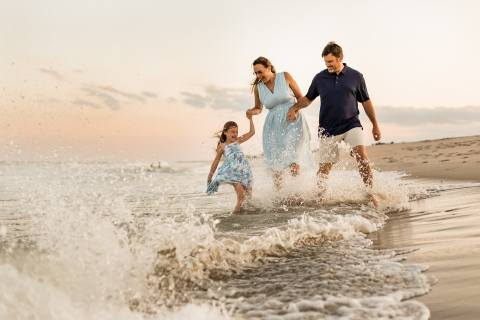 Une photo prise lors d'une séance photo de famille sur la côte du New Jersey montre une famille heureuse de trois personnes riant et se promenant sur la plage au bord de l'océan.